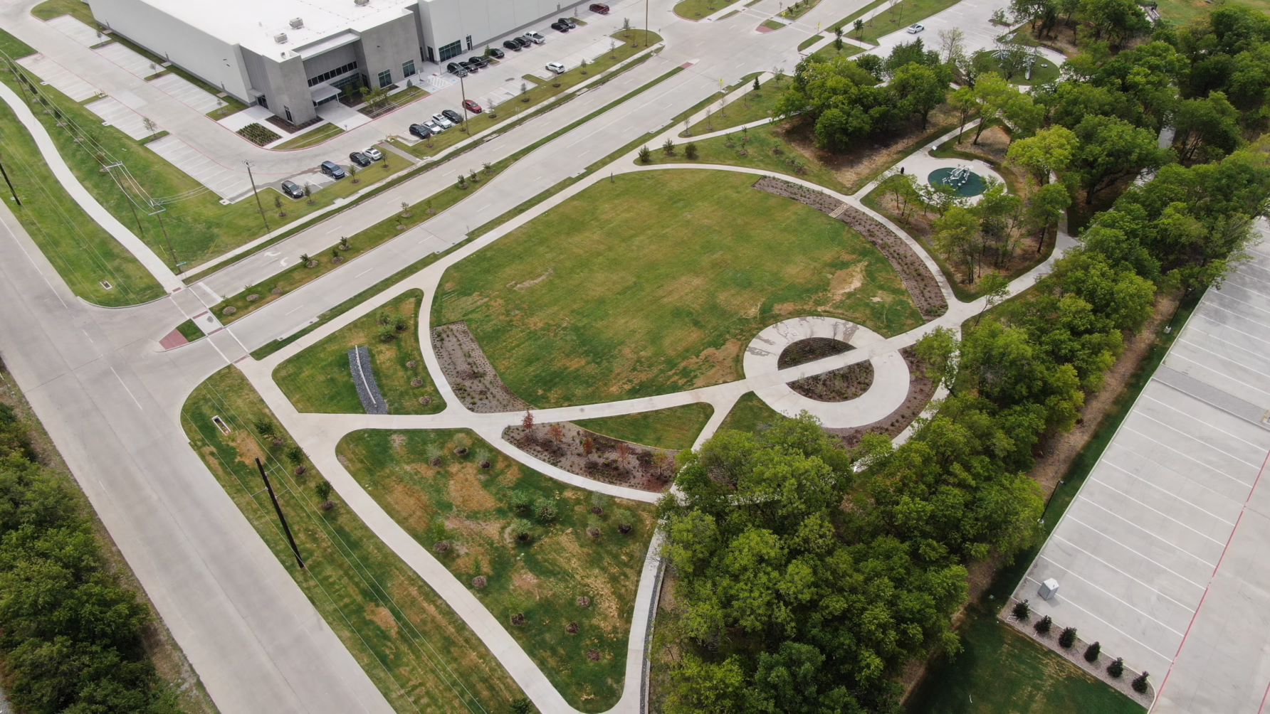 Aerial view of a landscaped park area with paths, trees, and adjacent industrial buildings.