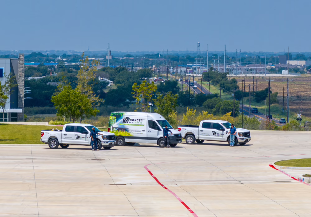 Landscape view with three utility vehicles and workers on a concrete surface, with trees and buildings in the background.