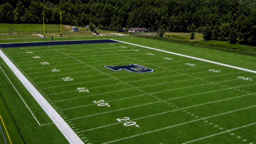 Aerial view of an empty football field with yard markings and goalposts surrounded by trees under a blue sky.