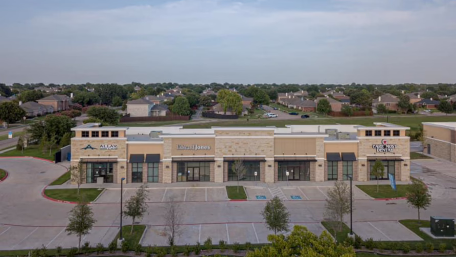 Aerial view of a shopping center with multiple storefronts and parking area on a clear day.