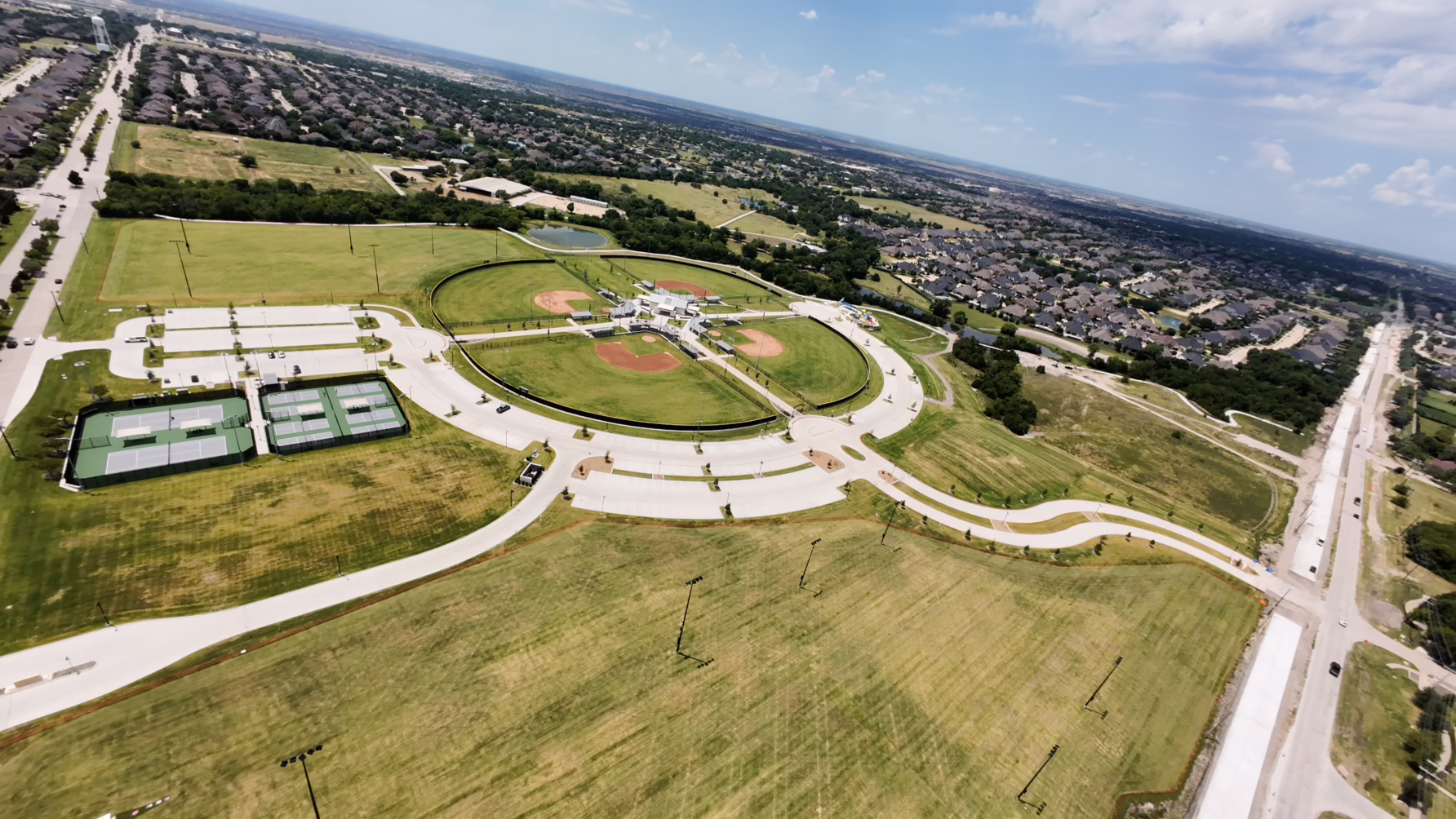 Aerial view of a park with baseball fields, tennis courts, and surrounding neighborhood on a sunny day.