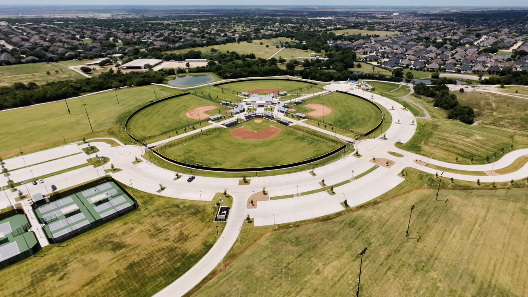 Aerial view of a sports complex with baseball fields, tennis courts, surrounding greenery, roads, and residential area.