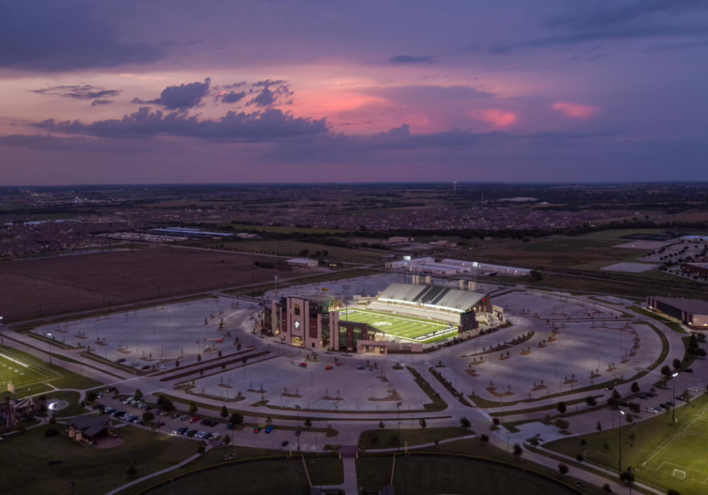 Aerial view of a stadium complex at sunset with illuminated field and expansive parking area, surrounded by vast open landscape.