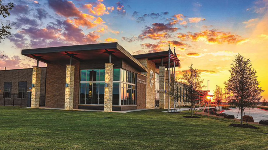 Modern building with large windows at sunset, surrounded by a grass lawn and trees, with a vibrant sky in the background.