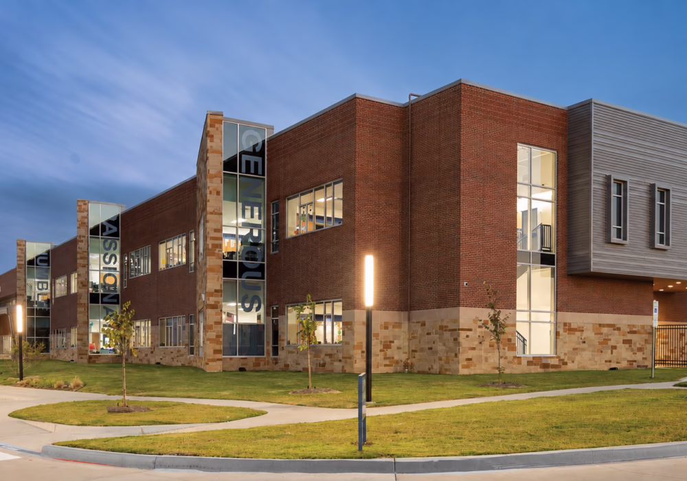 Modern two-story building with brick and stone exterior, large windows, surrounded by greenery and a sidewalk at dusk.