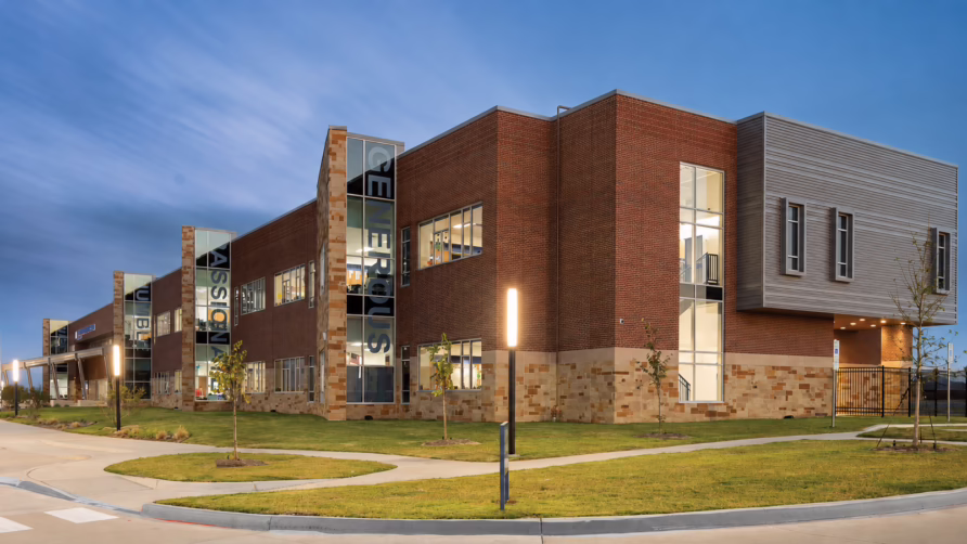 Modern two-story building with brick and stone exterior, large windows, surrounded by greenery and a sidewalk at dusk.